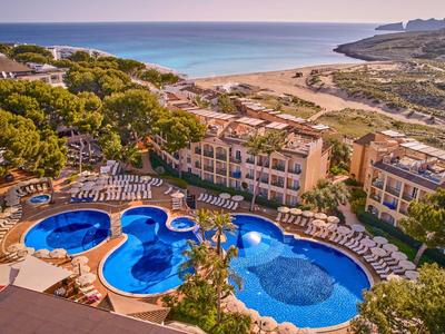 Landschaft mit Hotelanlage, drei verbundenen Pools, grünen Bäumen und Sandstrand am blauen Meer.