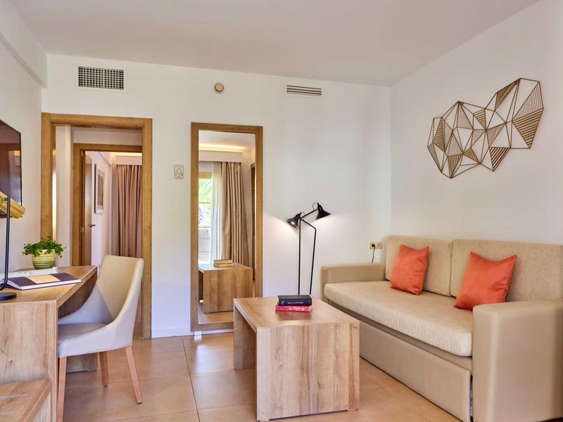 Světle zařízený hotelový pokoj s pohovkou, stolem, židlí a nástěnnou dekorací.