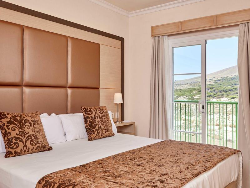 Světý hotelový pokoj s velkou postelí, dekoračními polštáři a balkonem s výhledem na hory.