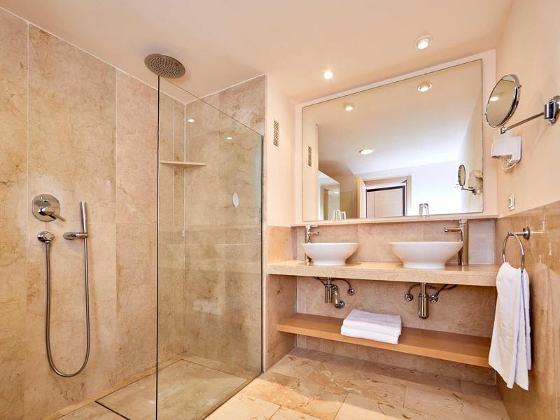 Moderní koupelna s bezbariérovým sprchovým koutem, dvojumyvadly a velkým zrcadlem v teplých odstínech.