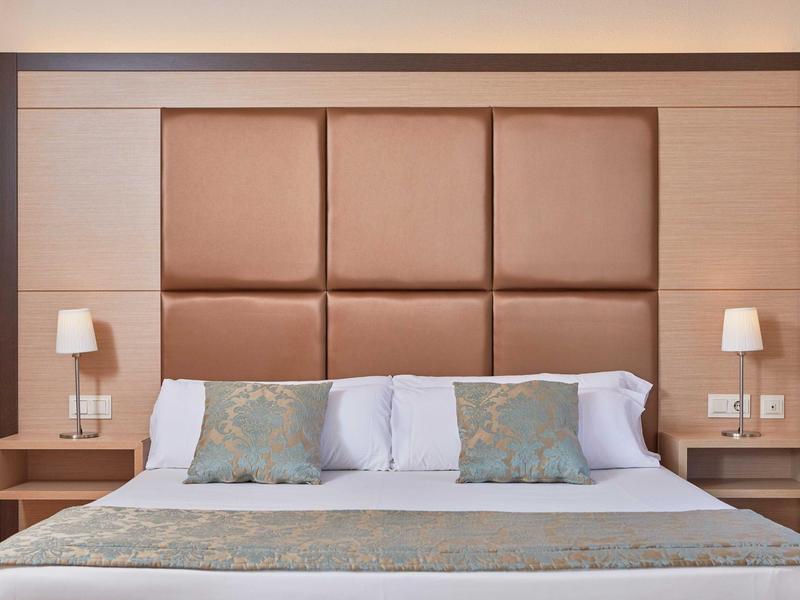 Moderně zařízený hotelový pokoj s velkou postelí, polstrovaným čelem a nočními stolky s lampami.