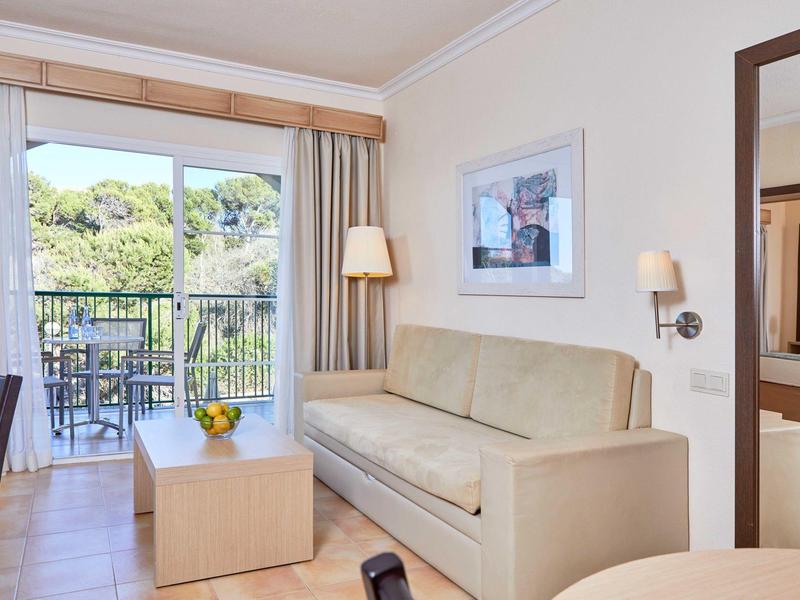 Světý obývací pokoj s béžovou pohovkou, dřevěným konferenčním stolkem a balkonem s výhledem na zelené stromy.