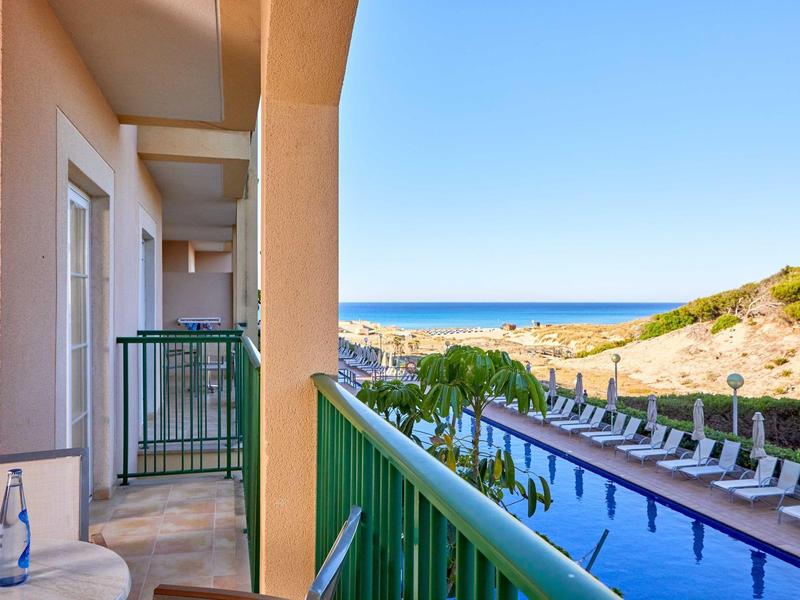 Balkon se zeleným zábradlím s výhledem na bazén a moře v pozadí