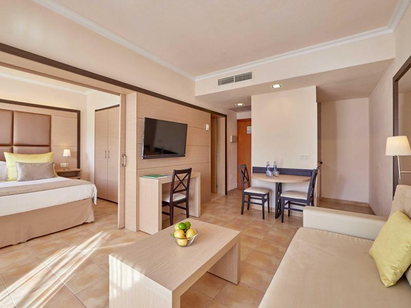 Světle hotelový pokoj s oddělenou ložnicí, pohovkou, televizí a jídelním stolem.