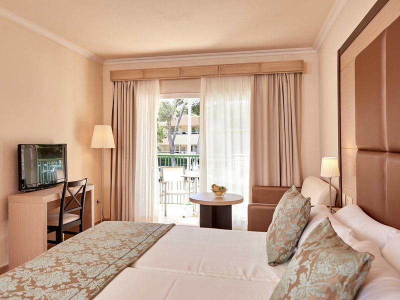Jasně zařízený hotelový pokoj s manželskou postelí, televizí a balkonem s výhledem do zahrady.