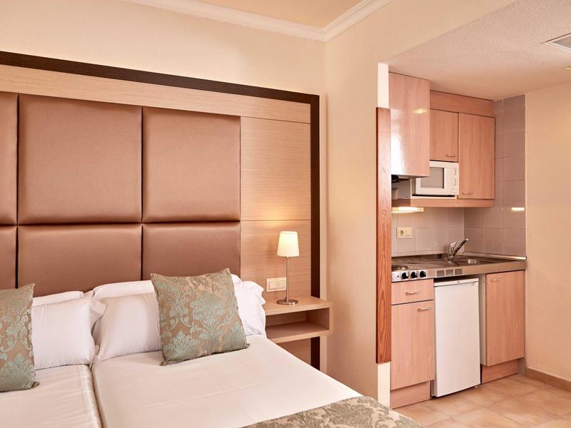 Hotelový pokoj s manželskou postelí, čalouněným čelem a malou kuchyňskou linkou vedle.