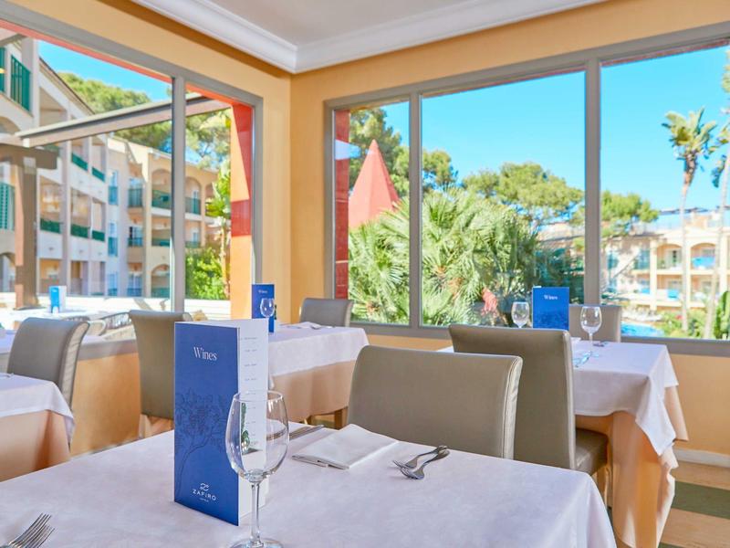 Světla jídelna s stoly, židlemi a velkými okny s výhledem na palmy.