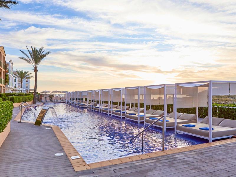 Moderní hotelový bazén s lehátky a pergolami při západu slunce.