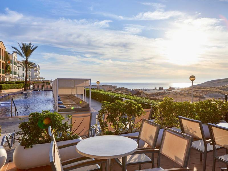 Hotelová terasa s stoly a židlemi, bazénem, rostlinami a výhledem na moře při západu slunce.