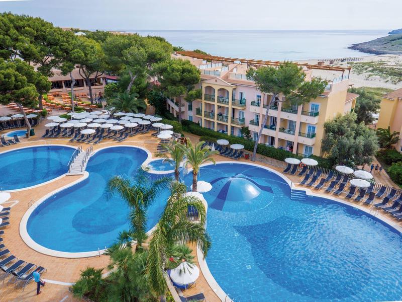 Velký hotelový bazén s lehátky, obklopený stromy s výhledem na moře v pozadí.