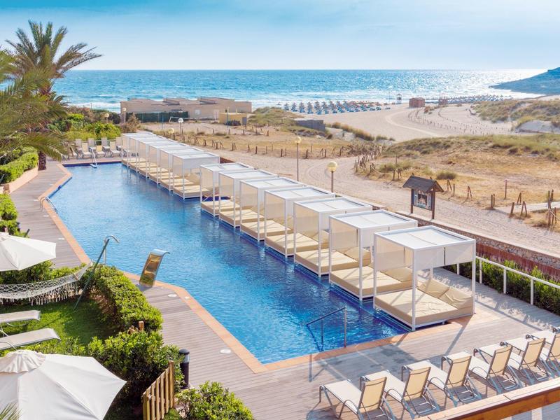 Moderní hotelový bazén s lehátky a výhledem na písečnou pláž a moře.