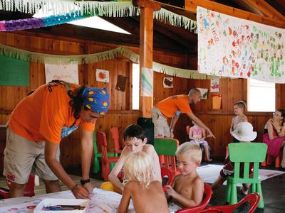 Bambini che dipingono a tavoli in una stanza con pannelli di legno, sorvegliati da due adulti.