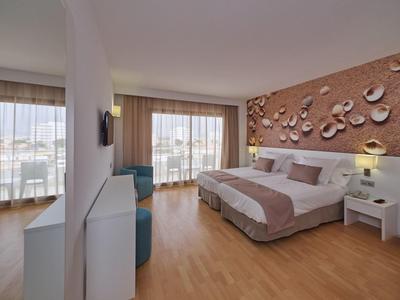 Habitación de hotel luminosa con cama doble, suelo de madera, balcón y pared decorativa con motivos de conchas.