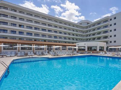 Complejo hotelero moderno con gran piscina exterior rectangular y tumbonas bajo cielo azul.