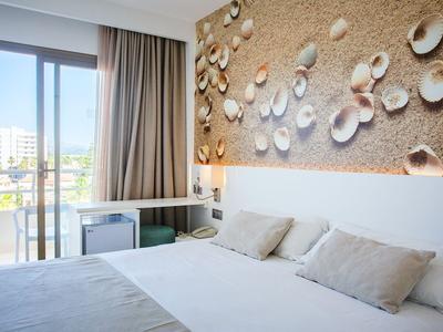 Camera d'hotel luminosa con letto matrimoniale, decorazioni con conchiglie e balcone con vista sulla città.