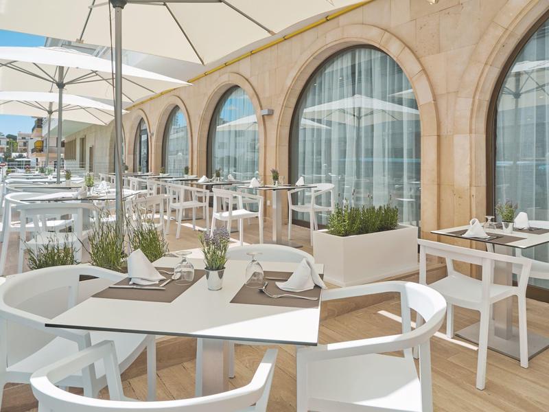 Terraza de restaurante al aire libre con mesas y sillas blancas junto a arcos de piedra.