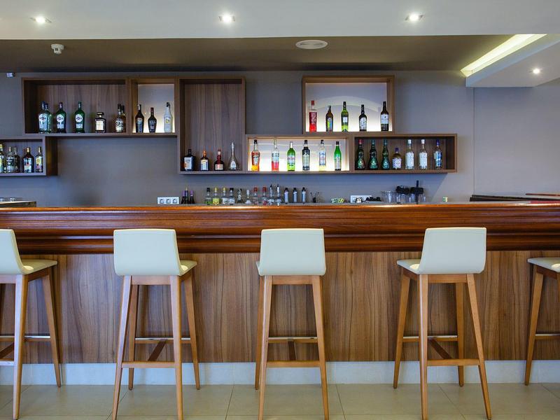 Área moderna de bar en hotel con cinco taburetes de madera y estantes llenos de bebidas.