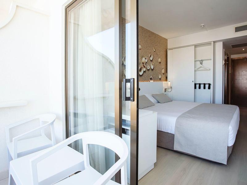 Vista a través de una puerta corrediza de vidrio abierta hacia una habitación de hotel luminosa con cama blanca y silla en el balcón.