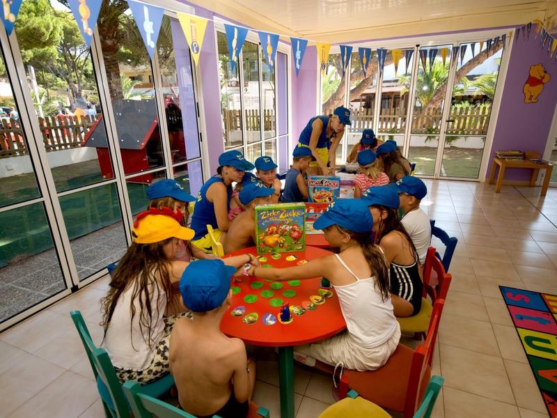 Bambini seduti a un tavolo rettangolare che giocano in una stanza colorata con grandi finestre.