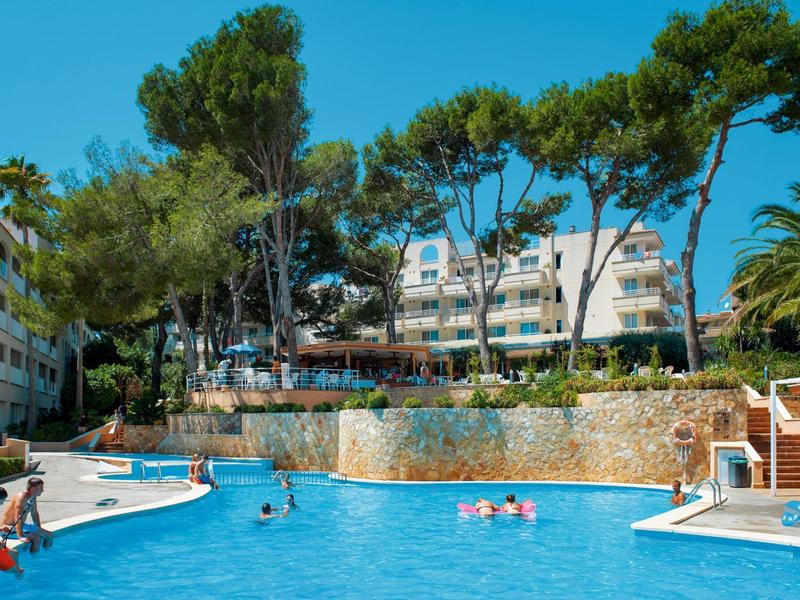 Area piscina dell'hotel con nuotatori, terrazza solarium e palme circostanti sotto un cielo sereno.