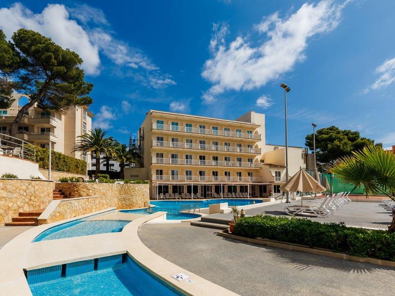 Complesso alberghiero moderno con piscina sotto un cielo azzurro chiaro circondato da alberi.