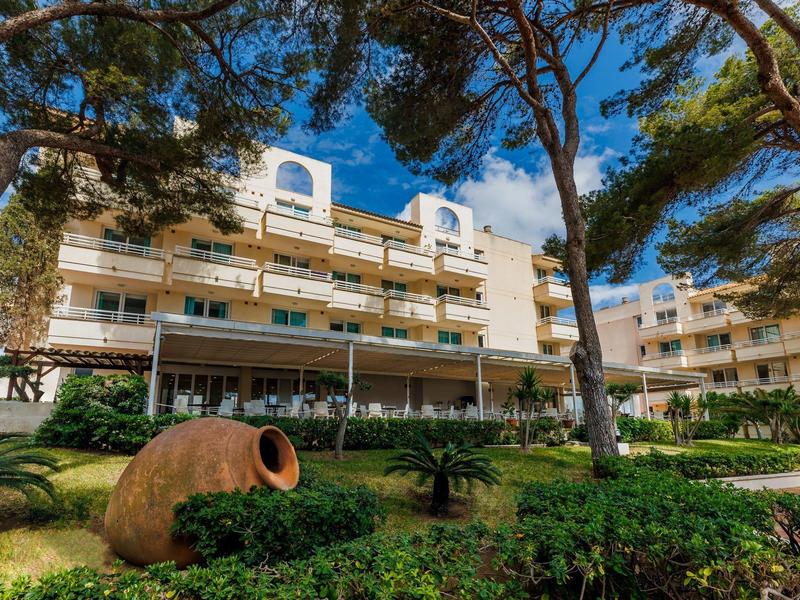 Hotel multistrato circondato da alberi e giardino curato sotto un cielo blu.