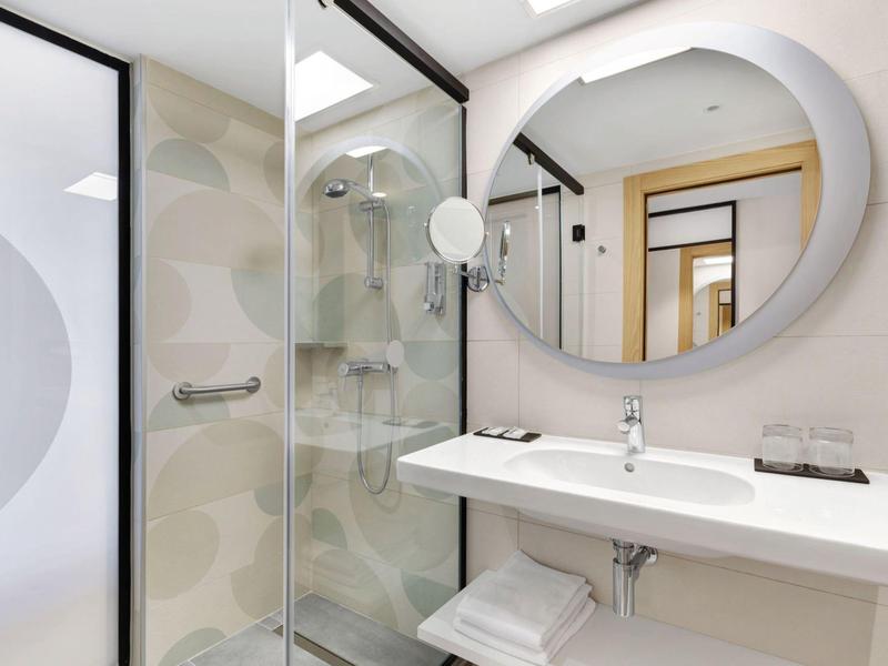 Baño moderno con espejo redondo, ducha de vidrio y lavabo blanco en una habitación de hotel.