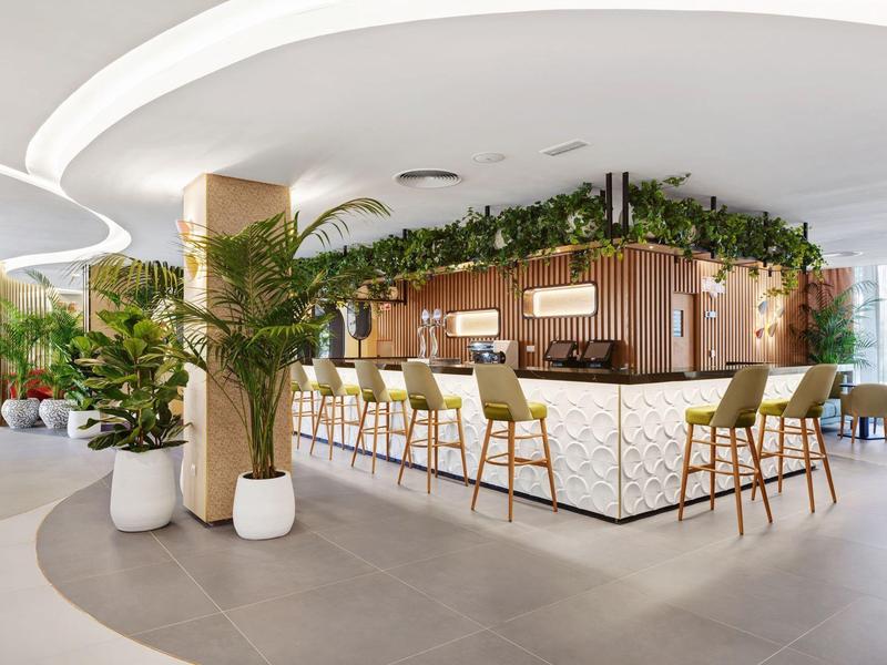 Bar moderna de hotel con plantas verdes y asientos claros bajo un techo blanco curvado.