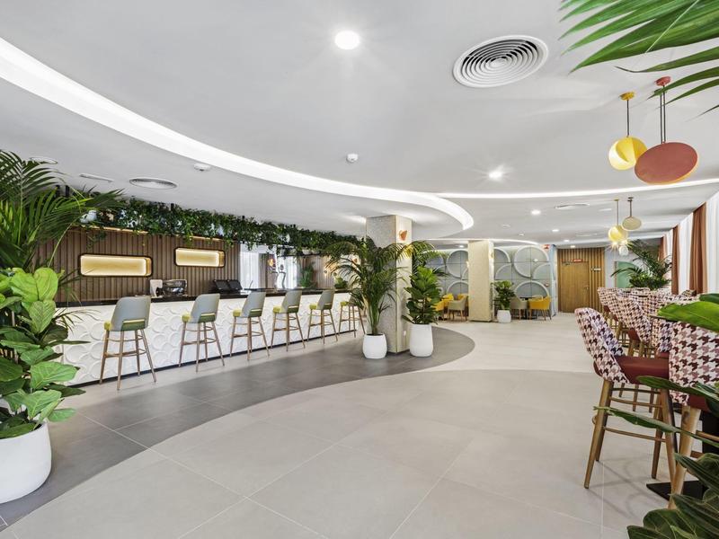 Área moderna de salón de hotel con bar, plantas verdes y asientos en un espacio luminoso.