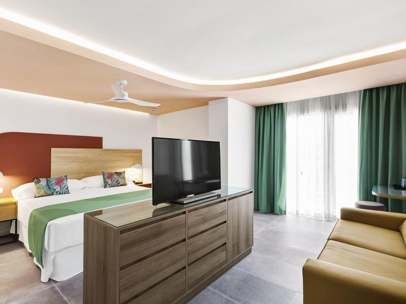 Habitación de hotel moderna con cama doble, televisor sobre cómoda y sofá junto a ventana con cortinas verdes.