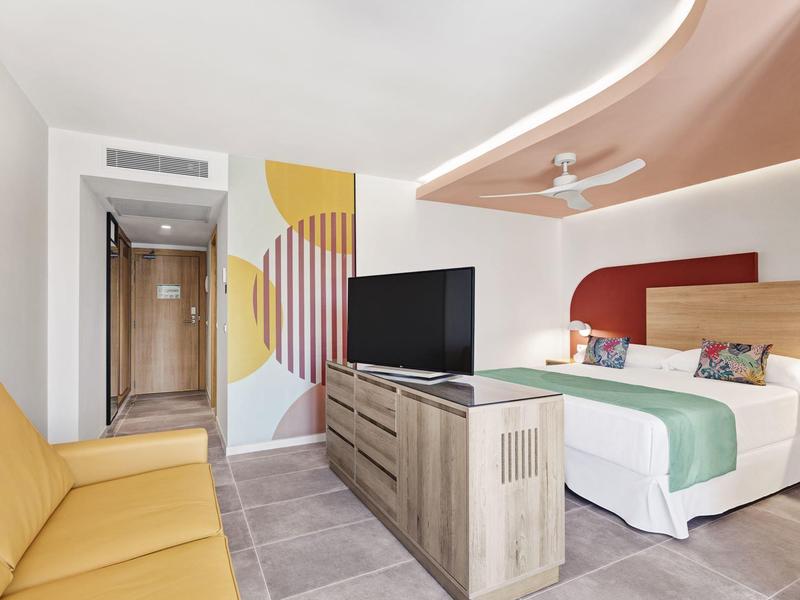 Habitación de hotel moderna con cama doble, sofá y televisor plano frente a una pared decorativa.