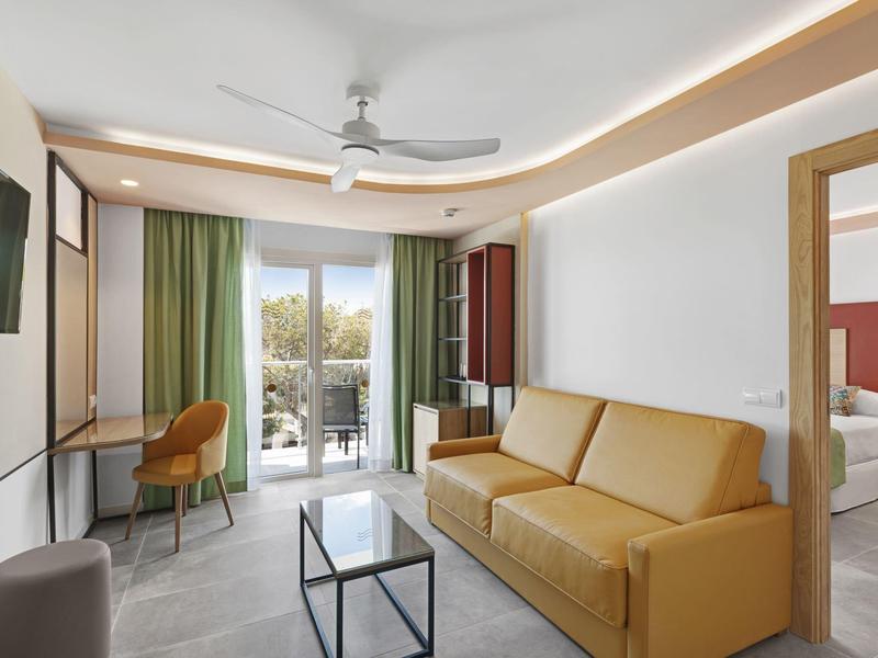 Sala luminosa con sofá amarillo, mesa de cristal, TV y acceso a balcón con cortinas verdes.