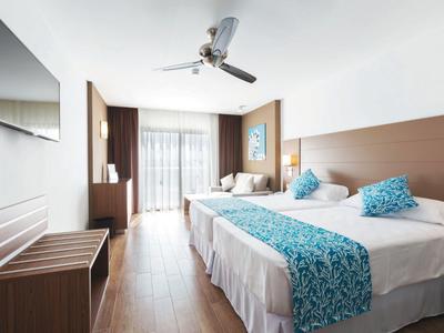 Helles Hotelzimmer mit Doppelbett, türkisen Akzenten, Deckenventilator und Balkon mit Vorhängen.