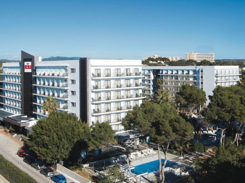 Moderní hotel s balkony, bazénem a stromy pod jasnou modrou oblohou.