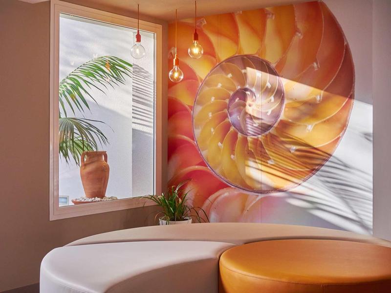 Nowoczesny pokój hotelowy z pomarańczowym okrągłym łóżkiem i dużą dekoracją ścienną muszli obok okna z rośliną.