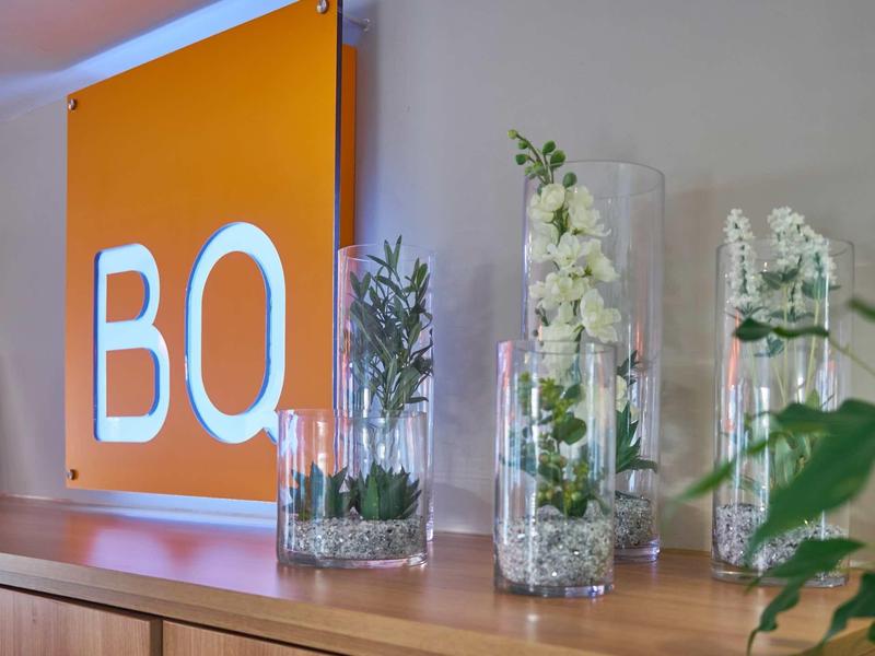 Dekoracyjne szklane wazony z roślinami przed pomarańczowym znakiem BQ na ścianie.