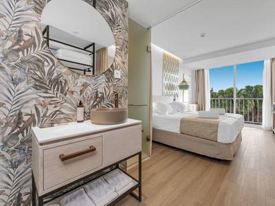Helles Schlafzimmer mit Doppelbett, großem Fenster, beige Vorhängen und modernem Waschbecken mit runder Spiegel.