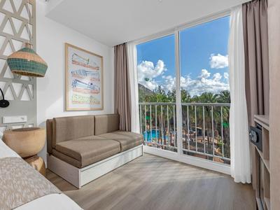 Helles Zimmer mit braunem Sofa, großem Fenster und Blick auf Pool und grüne Außenanlage.