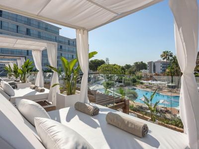 Luxuriöse Poolterrasse mit weißen Liegen, Vorhängen und tropischer Bepflanzung unter blauem Himmel.