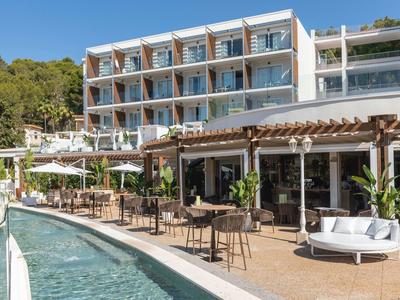 Modernes Hotel mit Glasbalkonen, Terrasse, Tischen, Stühlen und Pool vor bewaldetem Hügel.