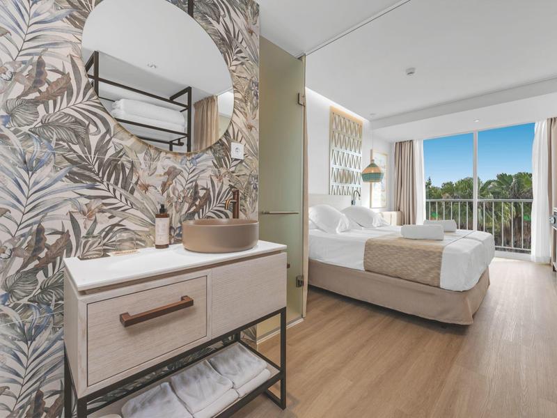 Moderně zařízený hotelový pokoj s velkou postelí, zrcadlem a balkonem s výhledem na zelenou přírodu.