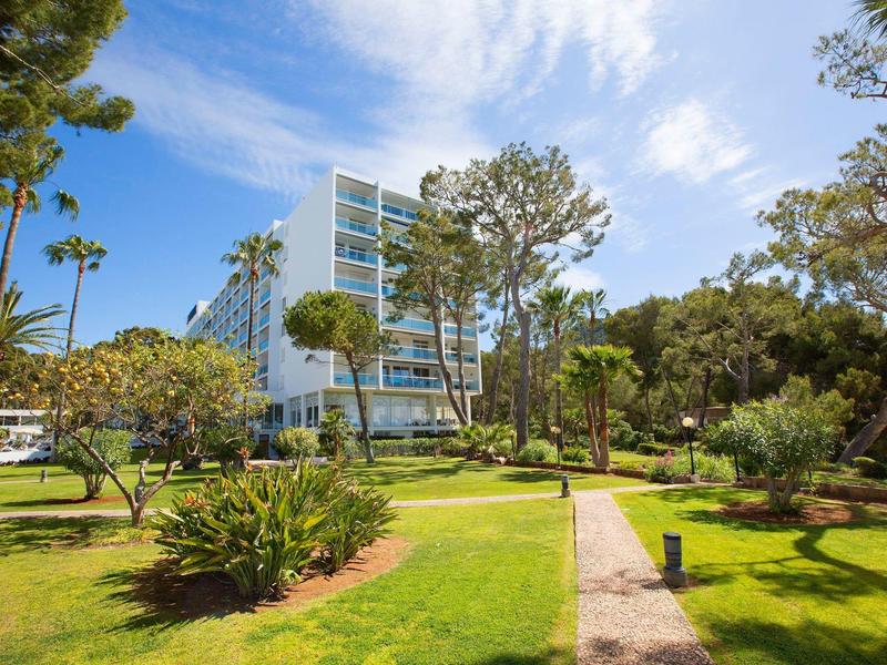 Moderní hotelová budova obklopená palmami a upravenou zahradou pod modrou oblohou.