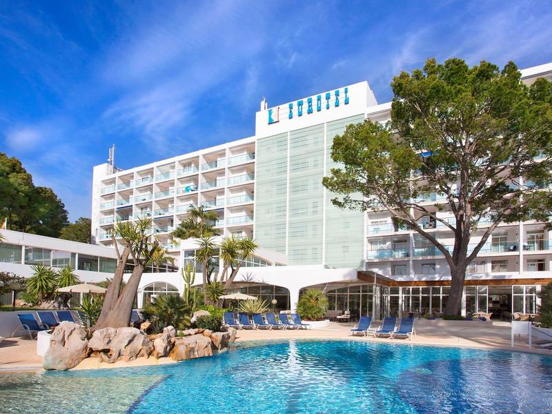 Moderní hotel s venkovním bazénem a palmami pod modrou oblohou.