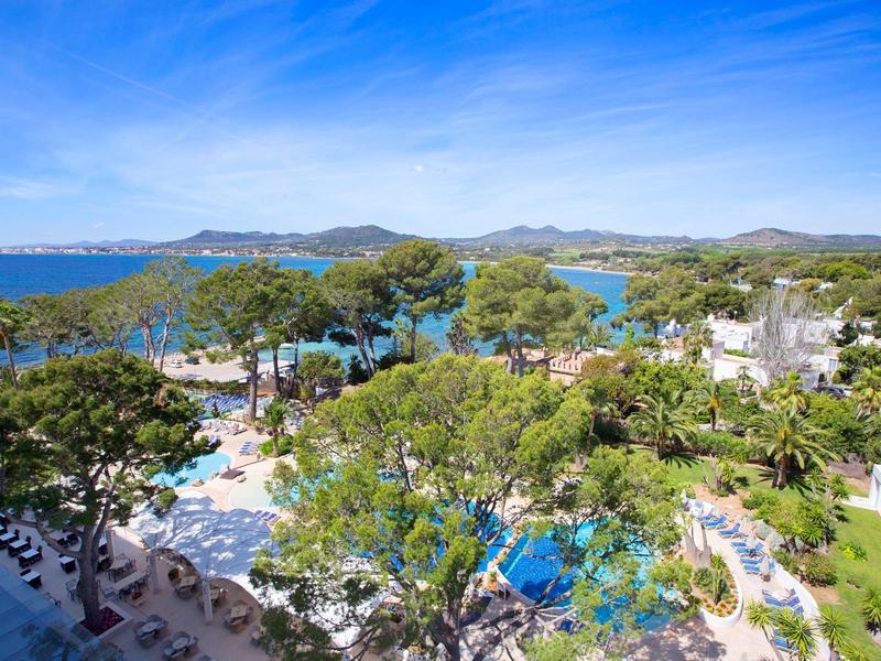 Pohled na hotelový bazén se stíněnými stromy a mořem v pozadí za jasné oblohy.