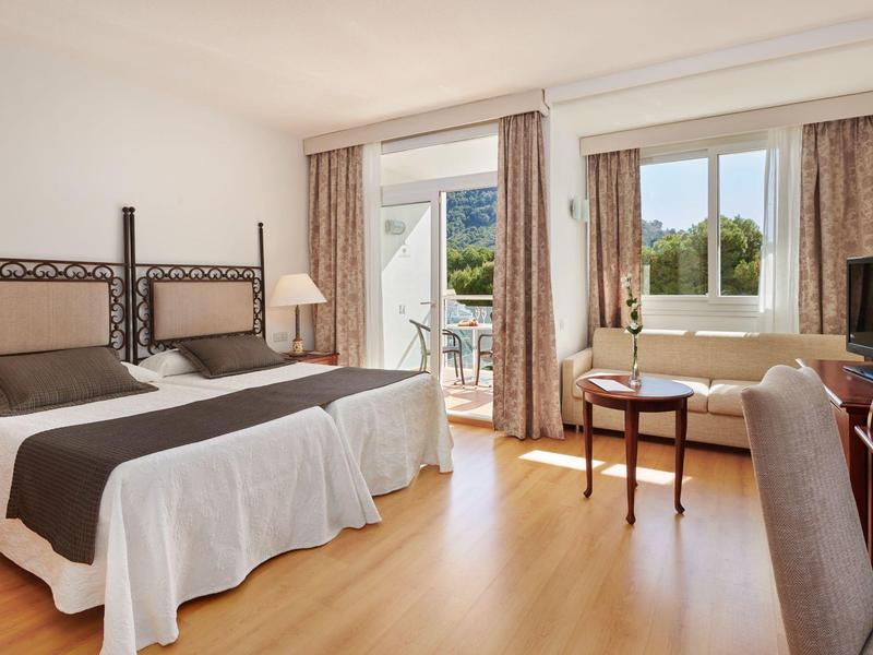 Světý hotelový pokoj se dvěma postelemi, dřevěnou podlahou a balkonem s výhledem na zelené stromy.