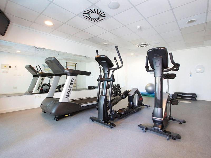 Moderní posilovna s běžeckým pásem, eliptickým trenažérem a stacionárním kolem na šedé podlaze.