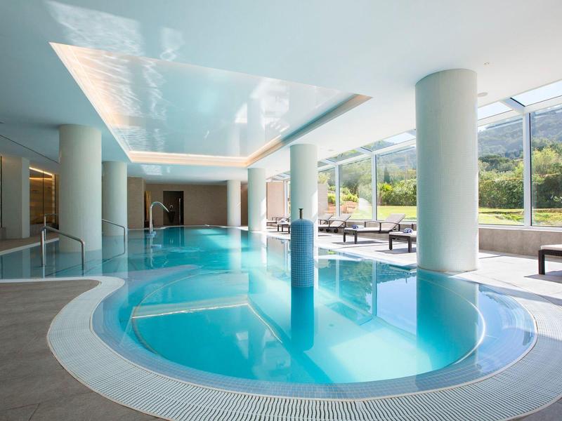 Vnitřní bazénová oblast se sloupy, posezením a velkými okny s výhledem na zeleň.