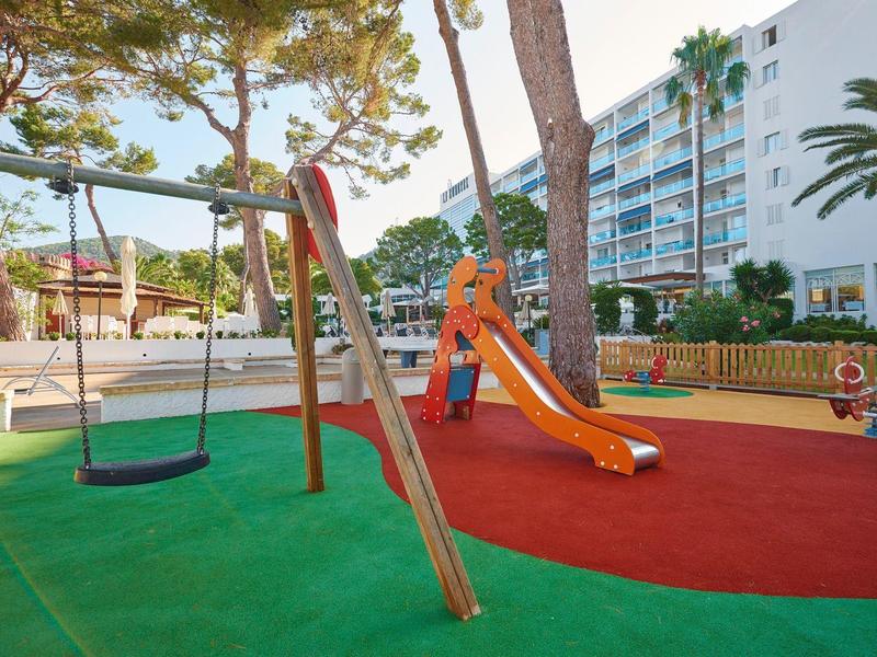 Hřiště s houpačkou, skluzavkou a měkkou podlahou před vícepodlažní hotelovou budovou.