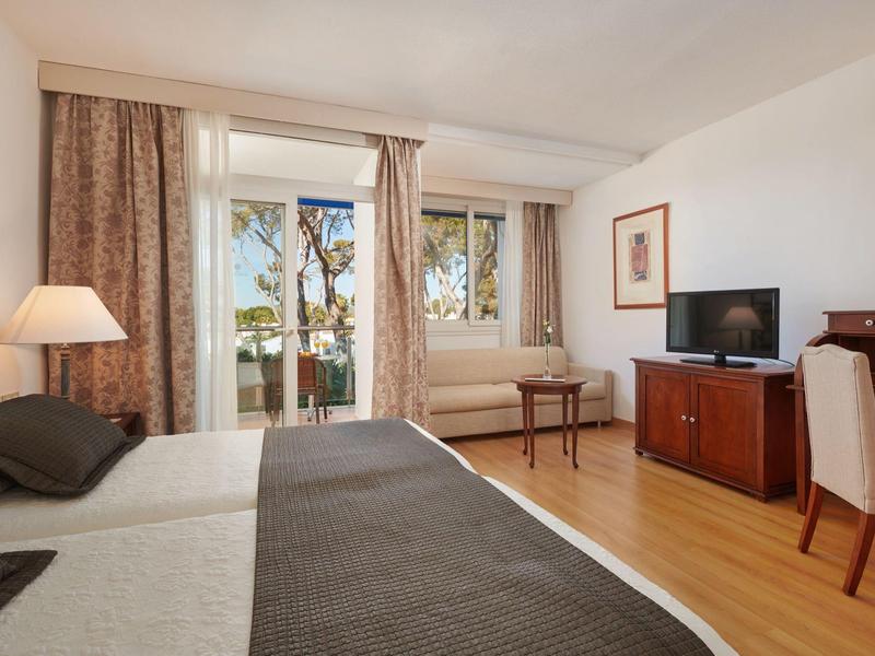 Světý hotelový pokoj s manželskou postelí, psacím stolem, televizí a přístupem na balkon s výhledem na palmy.