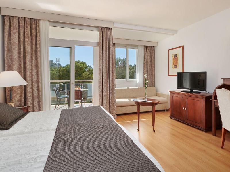 Světý hotelový pokoj s manželskou postelí, psacím stolem, televizí a balkonem s výhledem na zeleň.